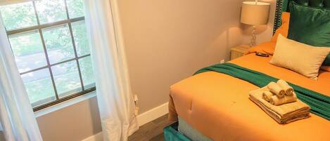 Een slaapkamer met een bed, gordijnen voor het raam, een bedlampje en uitzicht op groen.