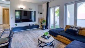 Ein modernes Wohnzimmer mit großem Flachbildfernseher, einer Couch, einem Couchtisch und einer Glastür, die zu einem Außenbereich führt.
