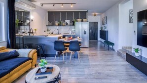 Ein modernes Wohnzimmer mit Sofa, Couchtisch und Fernseher. Im Hintergrund ist eine Küche mit Essbereich zu sehen.