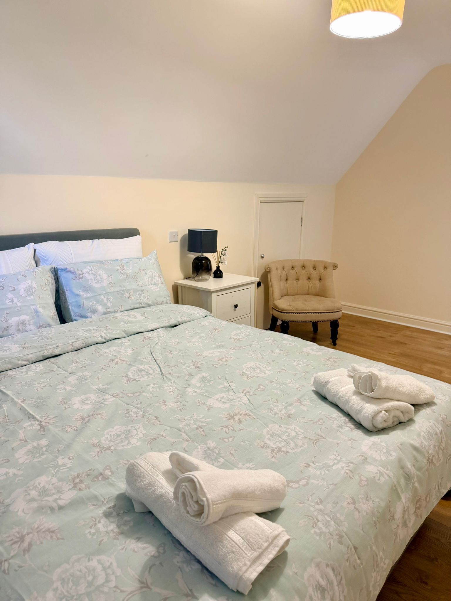 Een slaapkamer met een bed, nachtkastje, stoel en een deur.