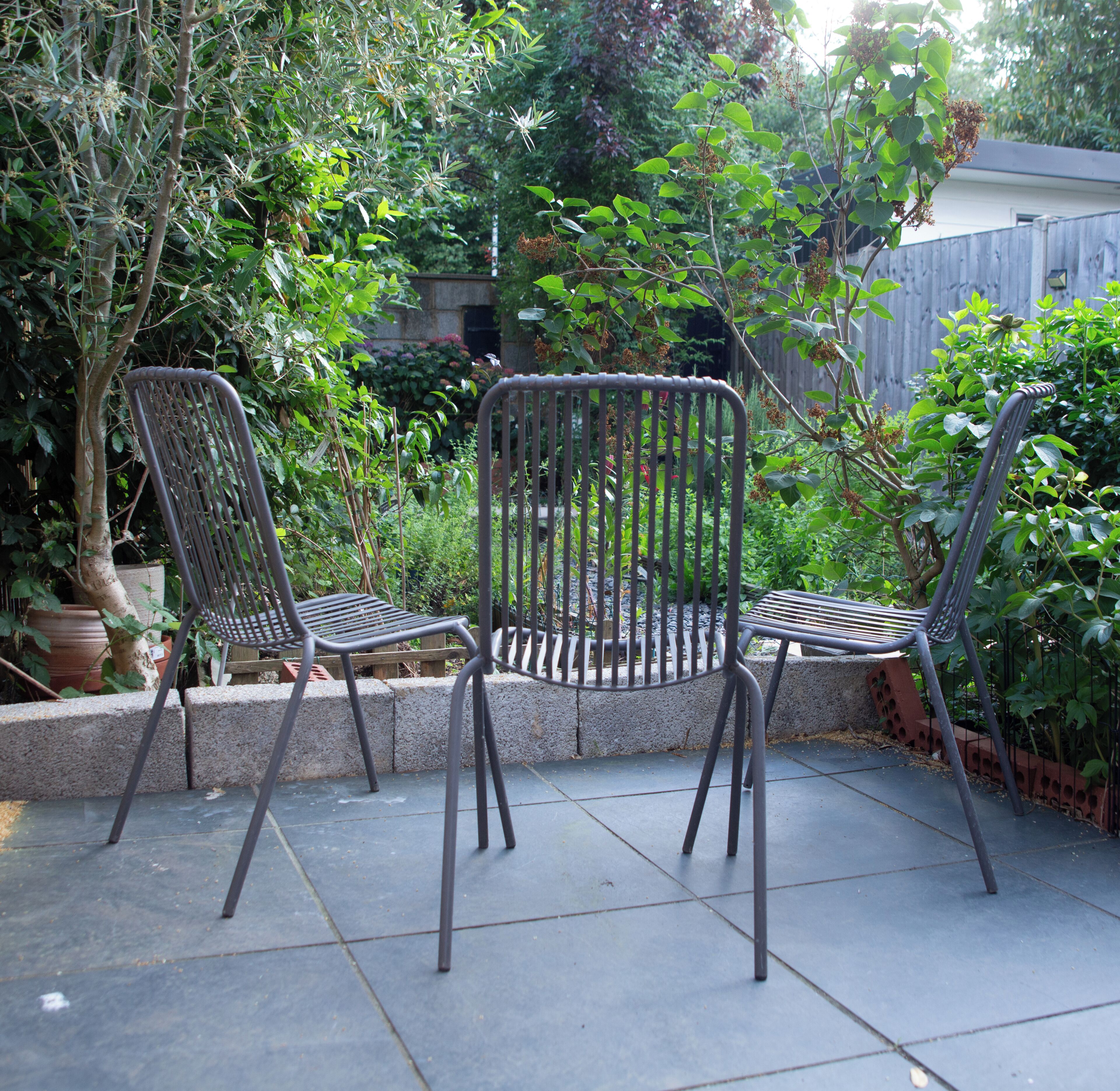 Two metal chairs with a metal grill backrest on a patio with a concrete border, surrounded by greenery and a wooden fence.