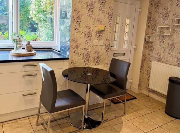 Une cuisine avec une table ronde, deux chaises, une fenêtre donnant sur un paysage verdoyant et une porte recouverte d’un papier peint à motifs floraux.