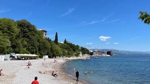 Een strand met mensen die zonnebaden en zwemmen, een heldere lucht en gebouwen op de achtergrond.