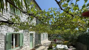 Un edificio de piedra con contraventanas verdes, un patio con mesa y sillas, y un jardín.