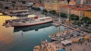 Un paisaje urbano frente al agua, con un gran crucero amarrado en un puerto deportivo, rodeado de edificios y un cielo despejado.