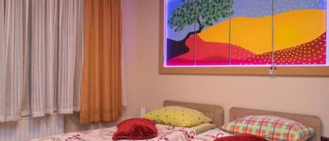 Quarto com cama, almofadas, janela e cortinas, e uma pintura colorida.