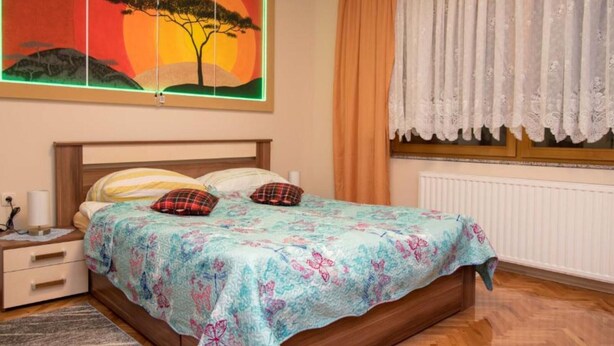 Un dormitorio con cama, mesita de noche, ventana con cortinas y un cuadro colgado en la pared.