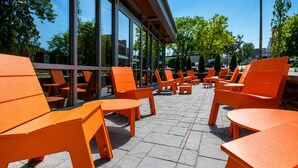 Ein Außenbereich mit orangefarbenen Stühlen und Tischen, großen Fenstern und einem Boden aus Steinplatten.