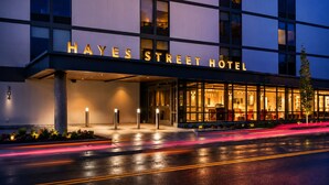 Ein modernes Hotel mit Glasfassade, der Schriftzug 'Hayes Street Hotel' und eine nasse Straße, die Lichter spiegelt.
