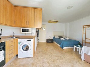 A compact hotel room with a kitchenette, a washing machine, a bed with a blue bedspread, and a bedside table with a lamp. - Ático A - Cartagena Centro histórico by Interhome (Cartagena)