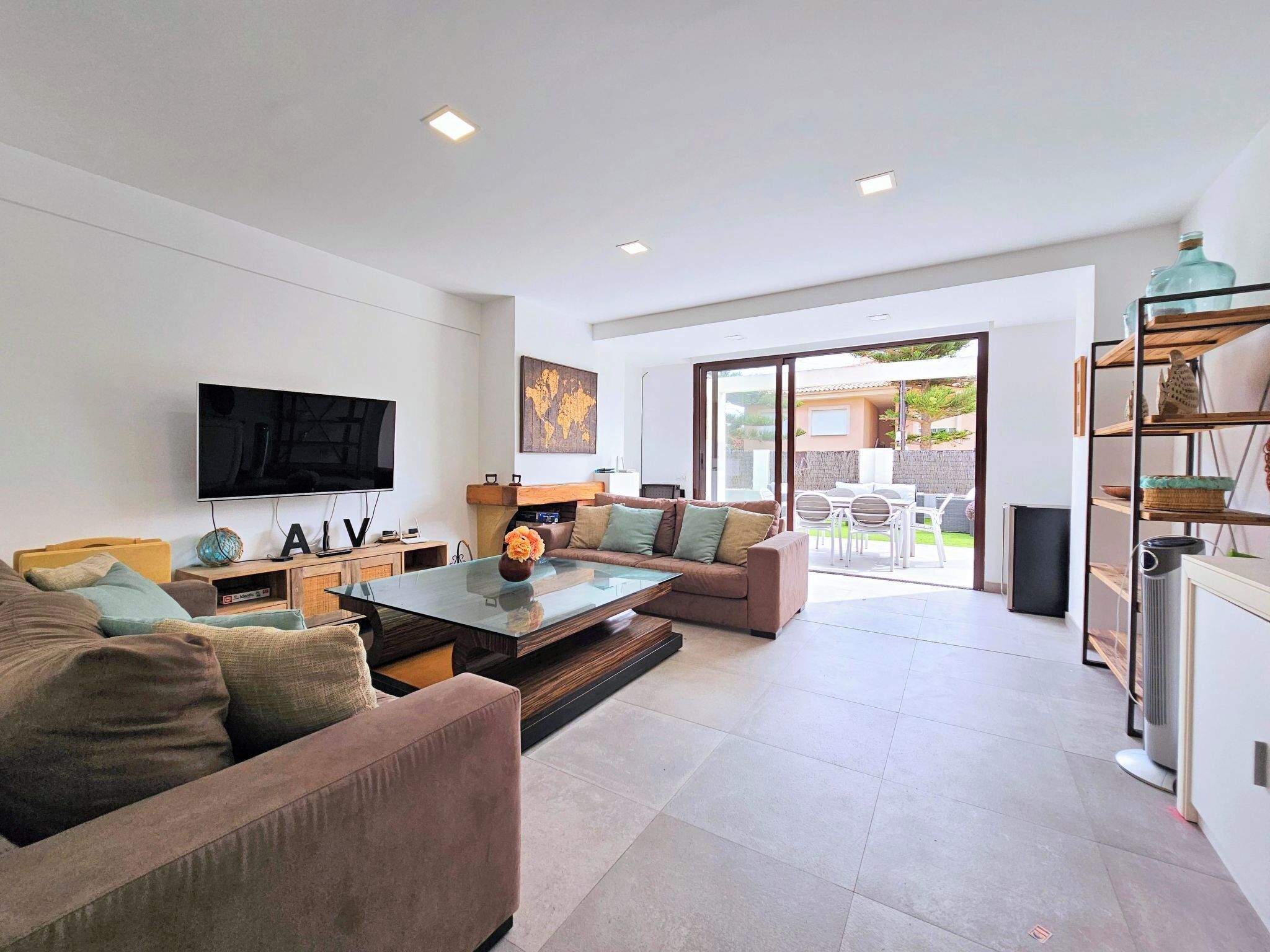 A modern living room with a flat-screen TV, a glass coffee table, and a sofa. There is a sliding glass door leading to an outdoor patio area.