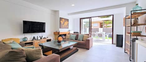 A modern living room with a flat-screen TV, a glass coffee table, and a sofa. There is a sliding glass door leading to an outdoor patio area.