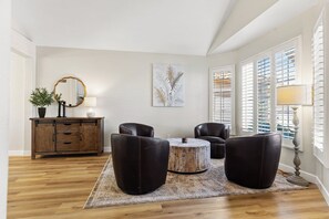 Ein Wohnzimmer mit Holzboden, einem runden Couchtisch, zwei Ledersesseln, einer Kommode mit Spiegel und einem großen Fenster mit Jalousien.