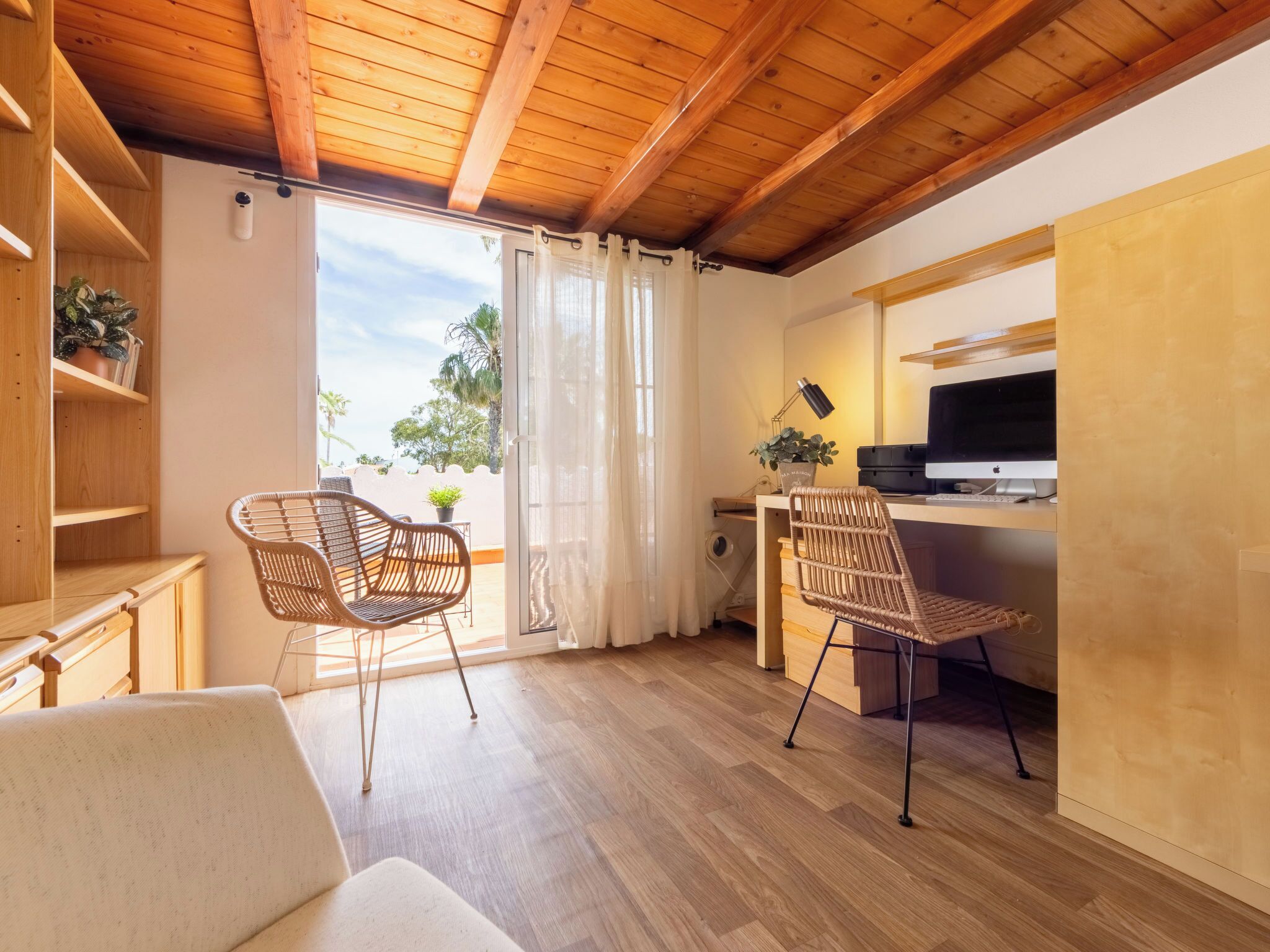 Una habitación con techo de madera, dos sillas de mimbre, un escritorio con una computadora, un televisor y una ventana con cortinas.