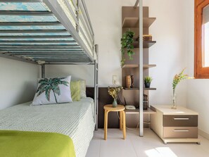 Ein Etagenbett mit grüner Decke, ein kleiner Holzschemel, ein Bücherregal mit Pflanzen und ein Nachttisch mit einer Vase.