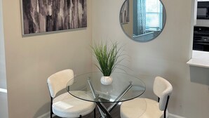 Ein Essbereich mit einem Glastisch und Stühlen, einem Blumentopf, einem großen abstrakten Gemälde und einem runden Spiegel an der Wand.