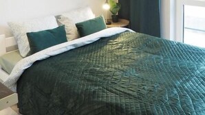 A bedroom with a bed, pillows, a nightstand, a plant, and a window with curtains. - Livin Apart A3 (Warszawa)