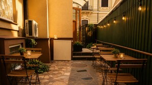 Un espace café en plein air avec un sol en pierre, des tables et des chaises en bois, des plantes en pot et des guirlandes lumineuses.