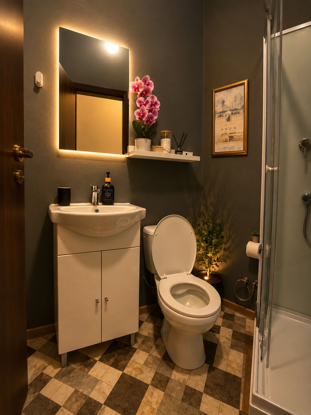 A bathroom with a white sink, toilet, and shower. There is a mirror above the sink, a framed picture on the wall, and a potted plant next to the toilet.