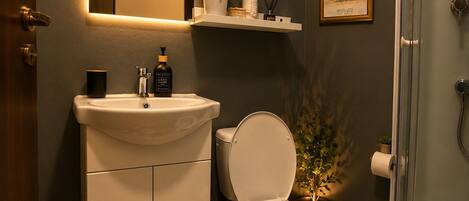 A bathroom with a white sink, toilet, and shower. There is a mirror above the sink, a framed picture on the wall, and a potted plant next to the toilet.