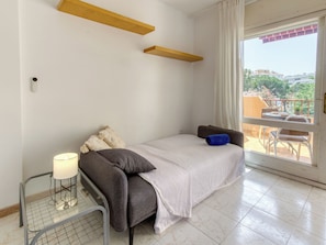 Ein Zimmer mit einem Bett, einem Nachttisch mit Lampe, einem Fenster mit Vorhängen und einem Balkon mit Gartenmöbeln.