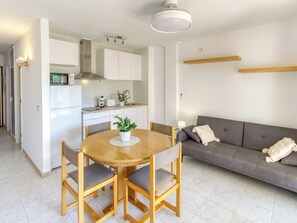 Eine moderne Küche mit weißen Schränken, einer eingebauten Mikrowelle und einem Herd. Ein Esstisch mit vier Stühlen und einer Topfpflanze steht dort. Ein graues Sofa mit Kissen lehnt an der Wand.