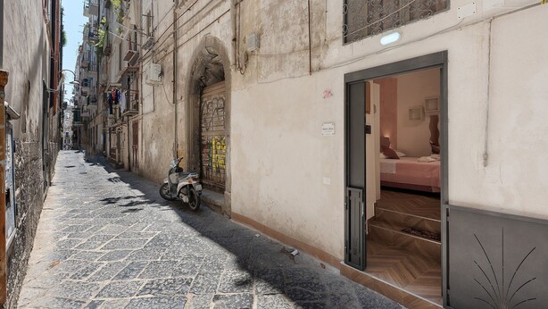 Uma rua estreita com uma scooter estacionada no acostamento, um edifício com porta e janela, e uma escadaria que leva a um andar superior.
