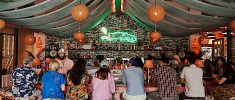 Um bar com clientes sentados em banquetas, um letreiro de neon e uma estante de livros atrás do balcão.
