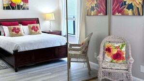 A bedroom with a bed, nightstand, lamp, wicker chair, and floral artwork. - Oceanfront 2/2 Condo w/ Panoramic Views on Del Monte Beach (Monterey)