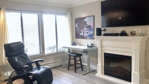 A living room with a fireplace, a mounted TV, a desk, and a leather chair. - Oceanfront 2/2 Condo w/ Panoramic Views on Del Monte Beach (Monterey)