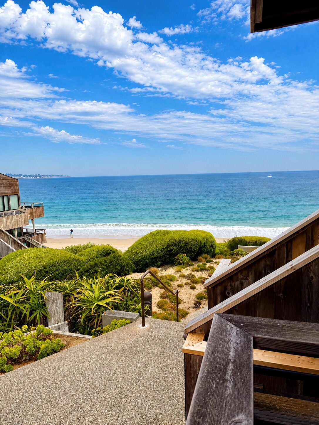 Una escalera de madera que baja a una playa, con vistas al océano y una costa lejana.