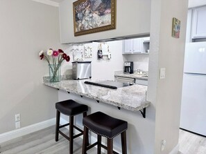 A kitchen with a marble countertop, two stools, a coffee maker, and a refrigerator. - Oceanfront 2/2 Condo w/ Panoramic Views on Del Monte Beach (Monterey)