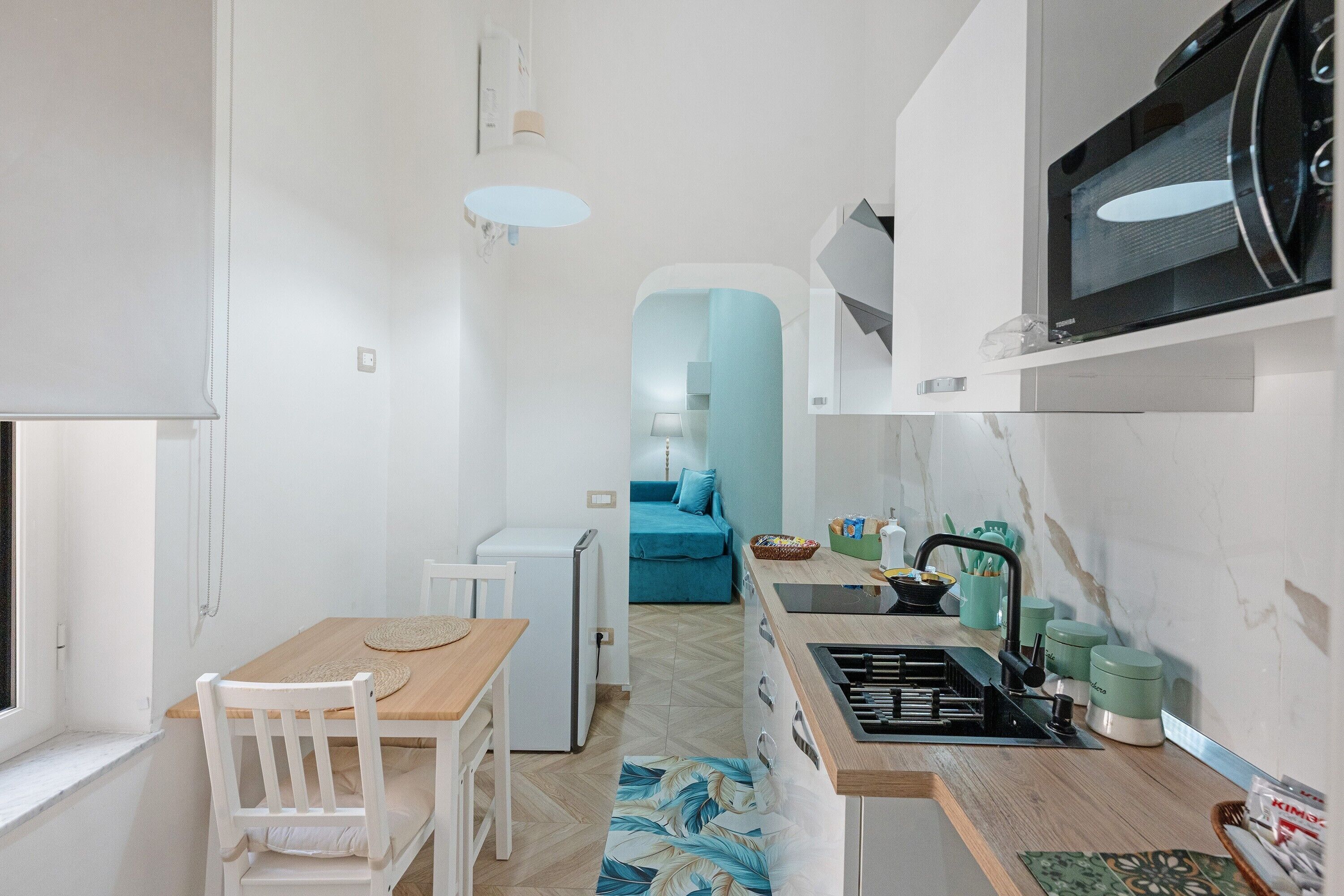 A compact kitchen with a microwave, sink, and small refrigerator. There is a dining table with chairs and a blue sofa in the background.