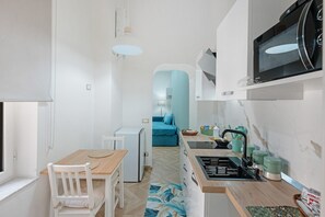 A compact kitchen with a microwave, sink, and small refrigerator. There is a dining table with chairs and a blue sofa in the background. - Apartment 'A casa re nonni' with Balcony, Wi-Fi and Air Conditioning (Naples)