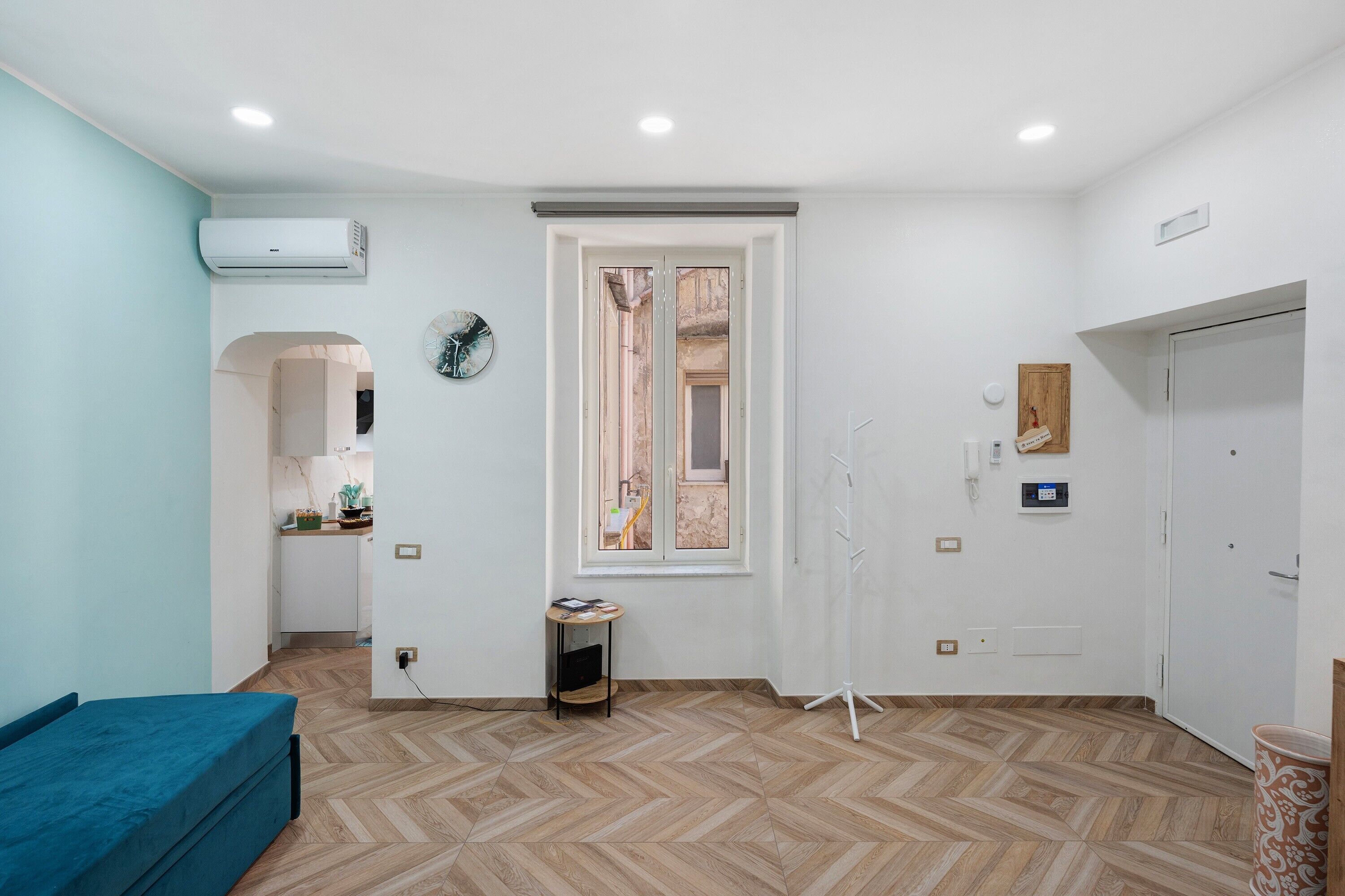 Une pièce dont le sol est recouvert d’un parquet à motifs en chevrons, avec un canapé bleu, une petite table sur laquelle sont posés divers objets, une horloge et une porte dotée d’un panneau en bois.