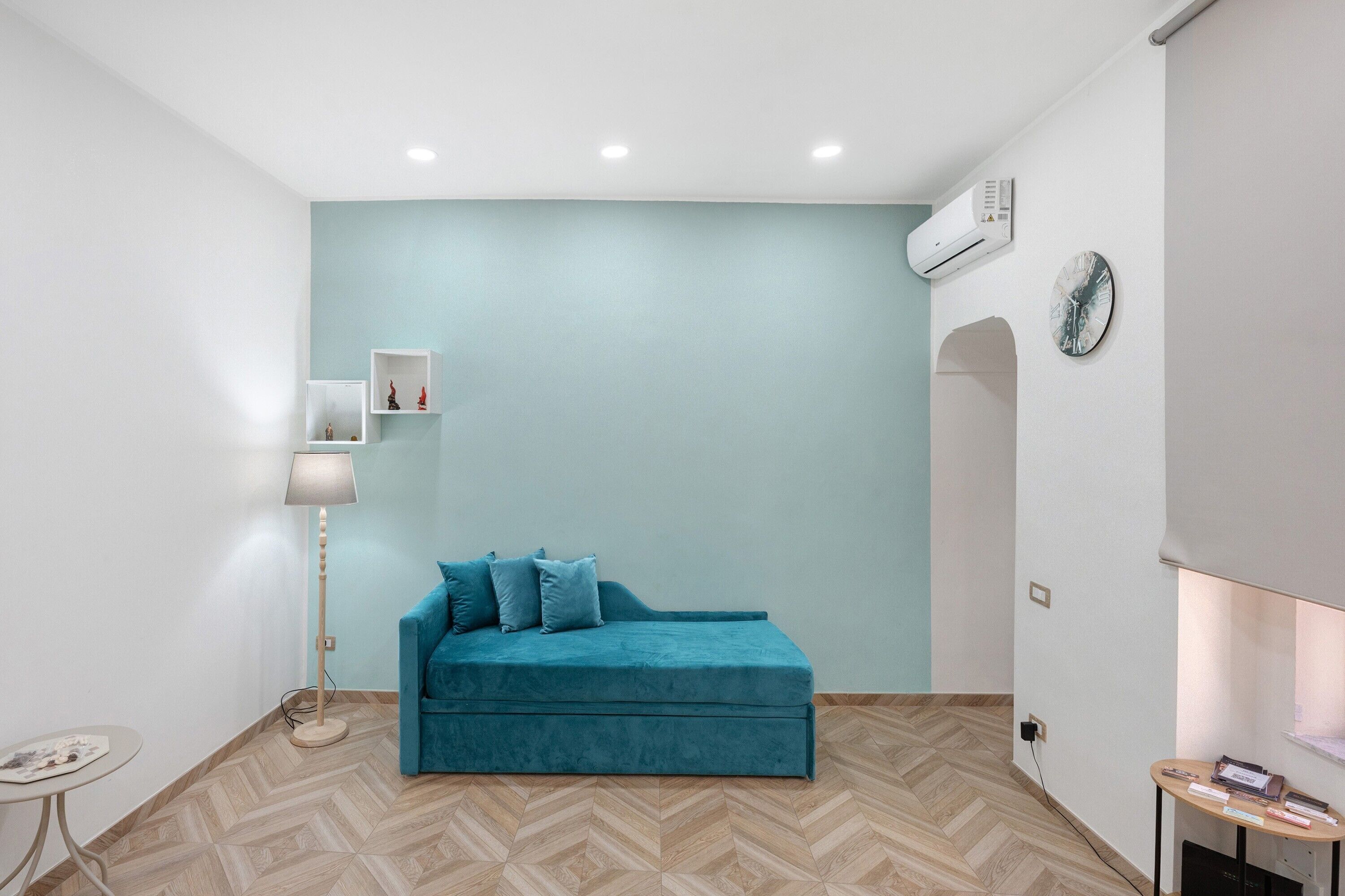 Ein Zimmer mit einem türkisfarbenen Sofa, einem kleinen Tisch, einer Stehlampe und einer Wanduhr.