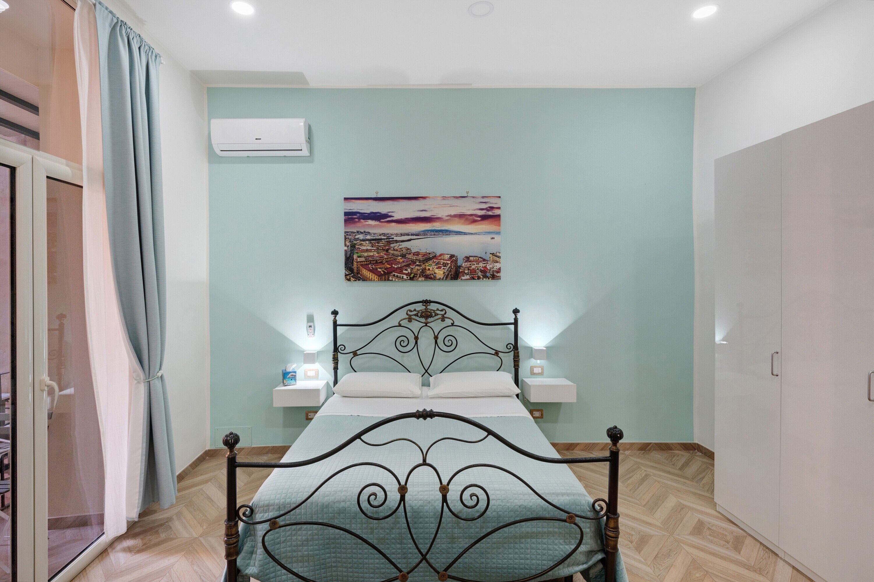 Un dormitorio con cama, mesita de noche, aire acondicionado instalado en la pared y un cuadro de un paisaje urbano.