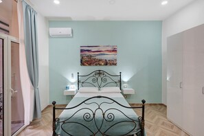 A bedroom with a bed, a nightstand, a wall-mounted air conditioner, and a painting of a cityscape. - Apartment 'A casa re nonni' with Balcony, Wi-Fi and Air Conditioning (Naples)