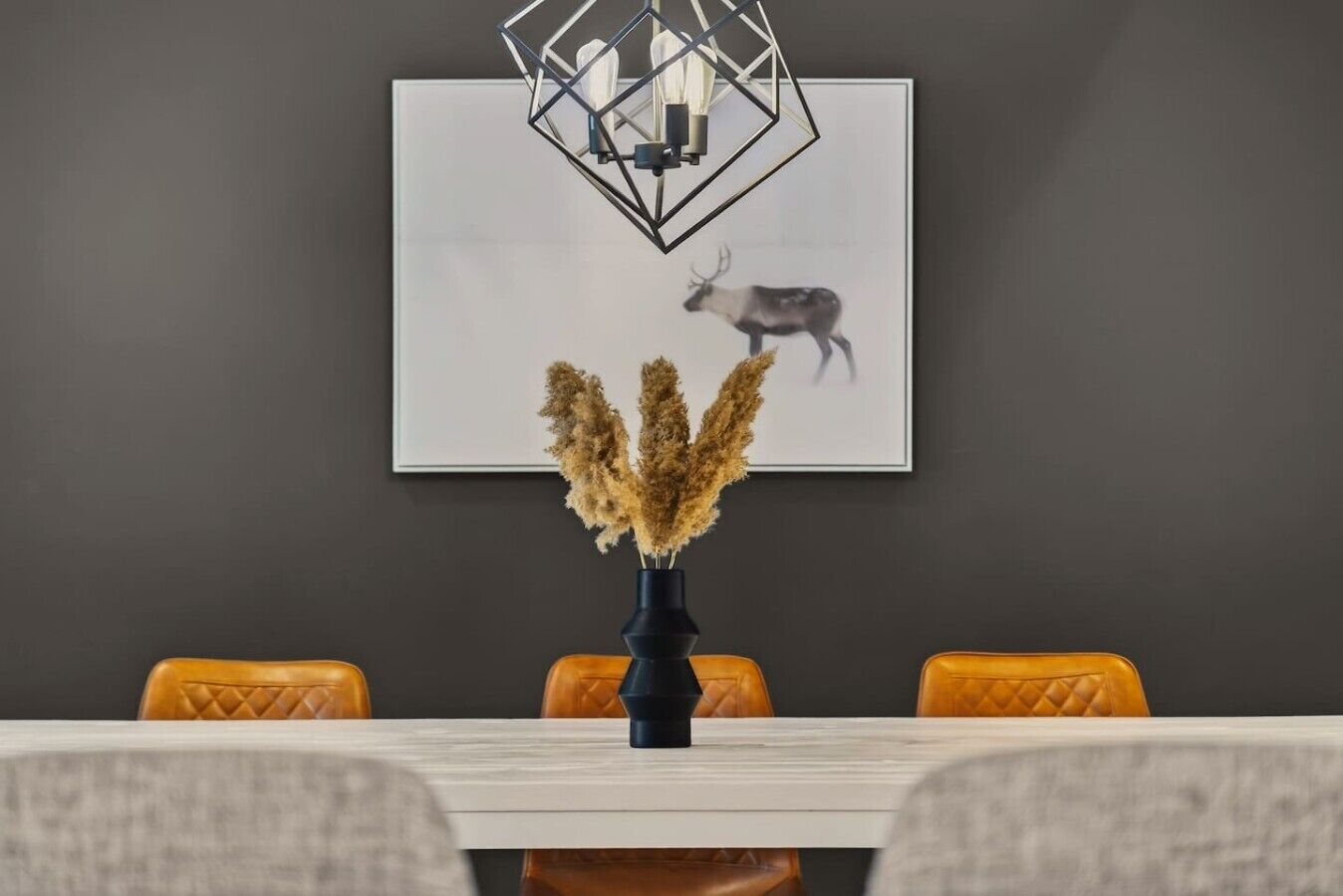 A dining area with a white table, a vase with dried plants, and a framed picture of a deer on the wall.