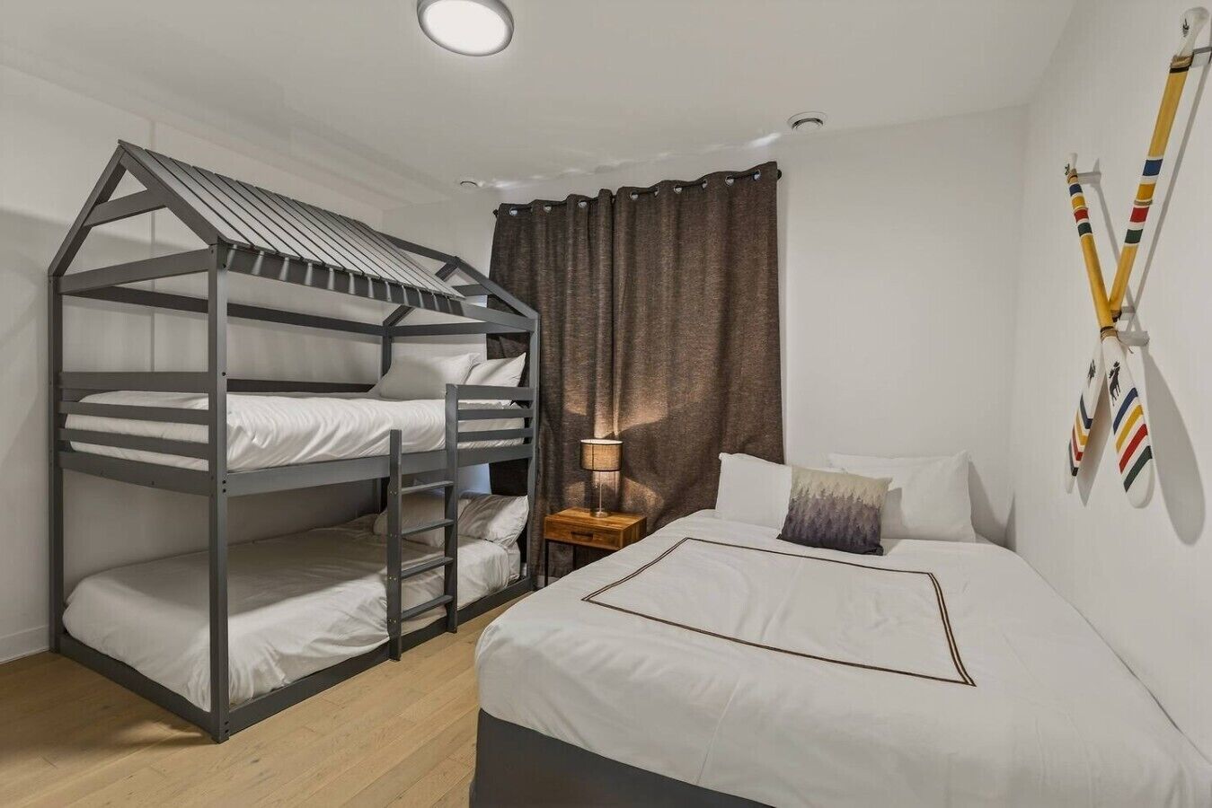 Una cama litera con escalera, una mesita de noche con lámpara, una cama con almohadas y una pared decorada con banderas deportivas.