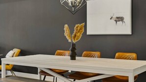 Un coin repas avec une table blanche, des chaises en cuir marron, un vase contenant des plumes d’herbe des pampas, un luminaire au design géométrique et un tableau encadré représentant un renne accroché au mur.