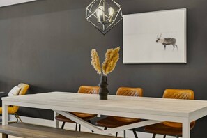 A dining area with a white table, brown leather chairs, a vase with pampas grass, a geometric light fixture, and a framed picture of a reindeer on the wall. - R&R at Verbier | Luxury Tremblant Retreat (Mont-Tremblant)