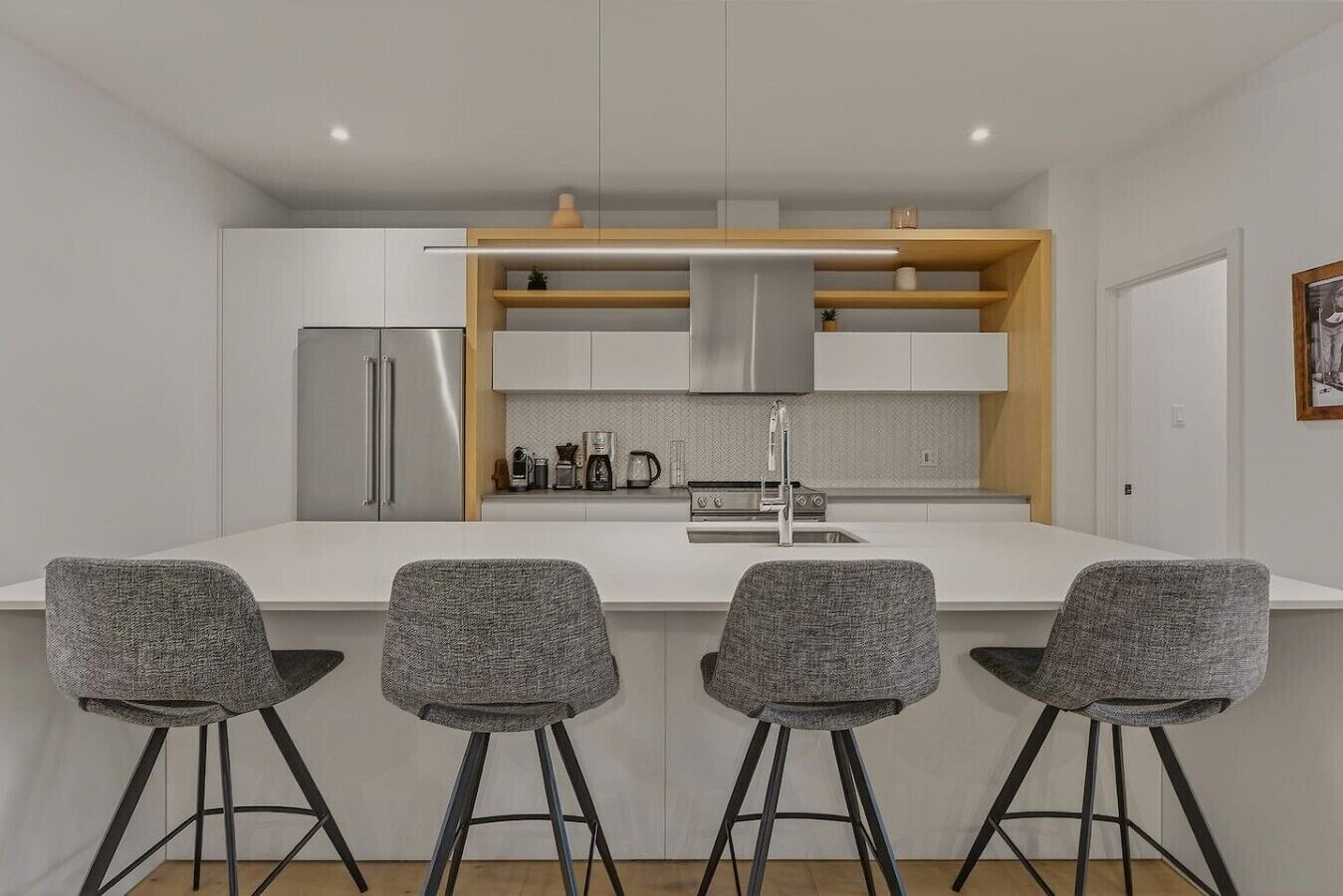Una cocina moderna con armarios blancos, un refrigerador de acero inoxidable, un horno empotrado y un estante de madera sobre la encimera. Hay tres taburetes grises con patas negras.