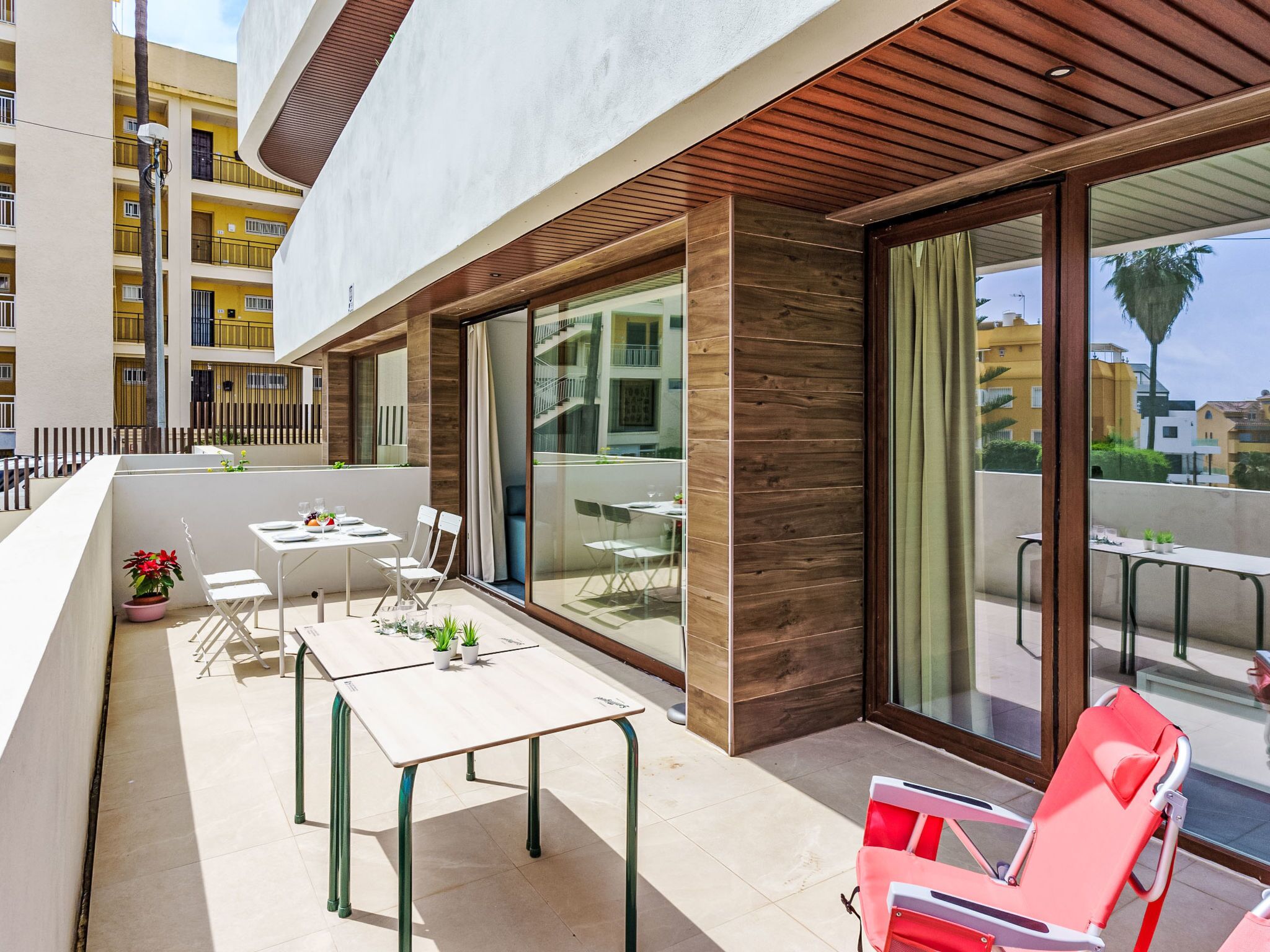 A modern outdoor patio with a table and chairs, a red lounge chair, and a view of neighboring buildings.
