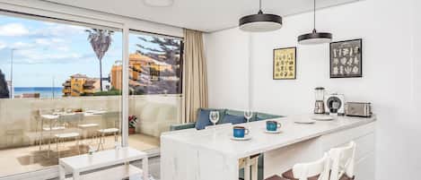 A modern kitchen with a marble countertop, white chairs, a dining table, and a view of the sea.