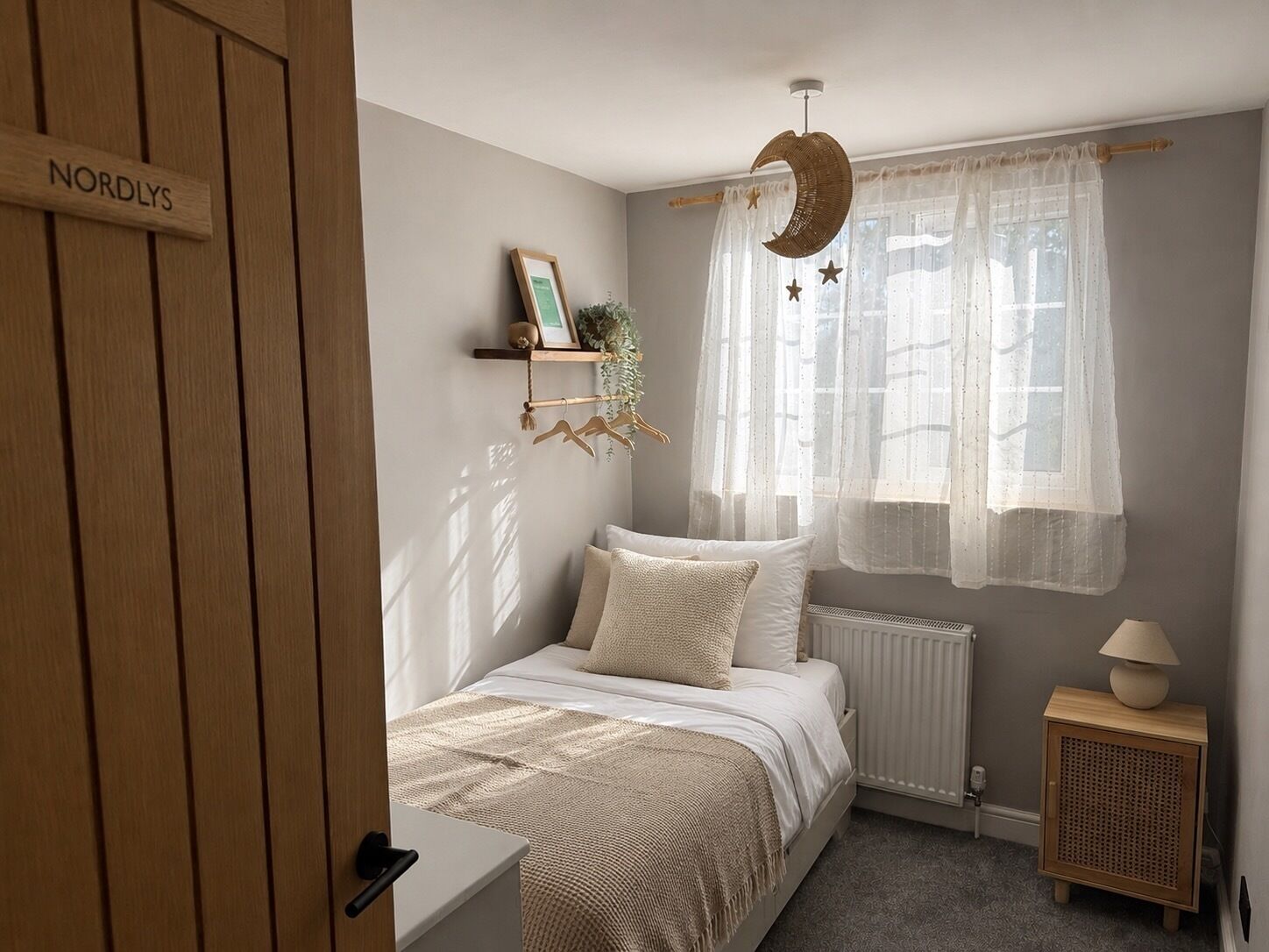 Un dormitorio con cama, una mesita de noche, cortinas translúcidas en la ventana, una lámpara colgante con forma de luna y un radiador.