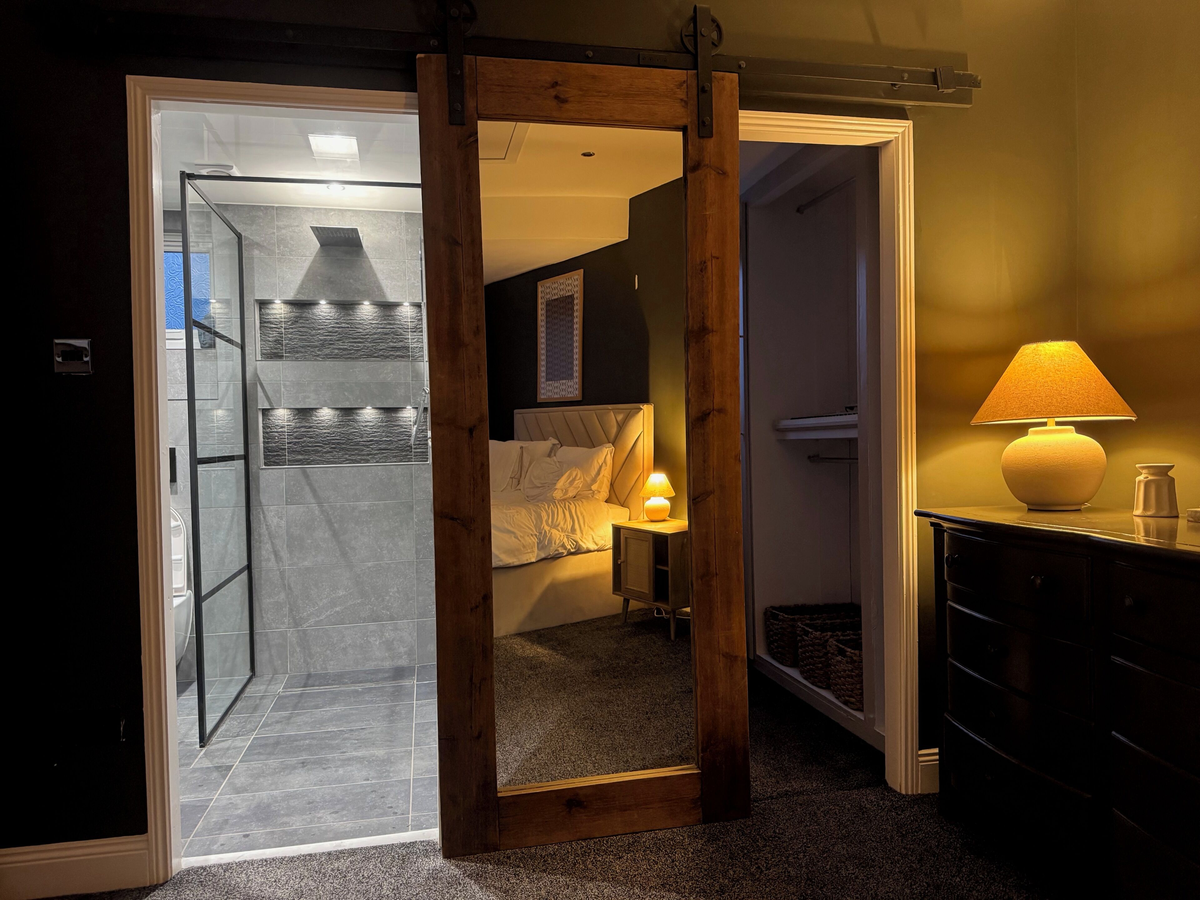 Una habitación con cama, una mesita de noche con lámpara, un marco de puerta de madera y vistas a un baño con ducha.