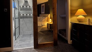 Une chambre avec un lit, une table de chevet avec une lampe, un cadre de porte en bois et une vue sur une salle de bain avec une douche.