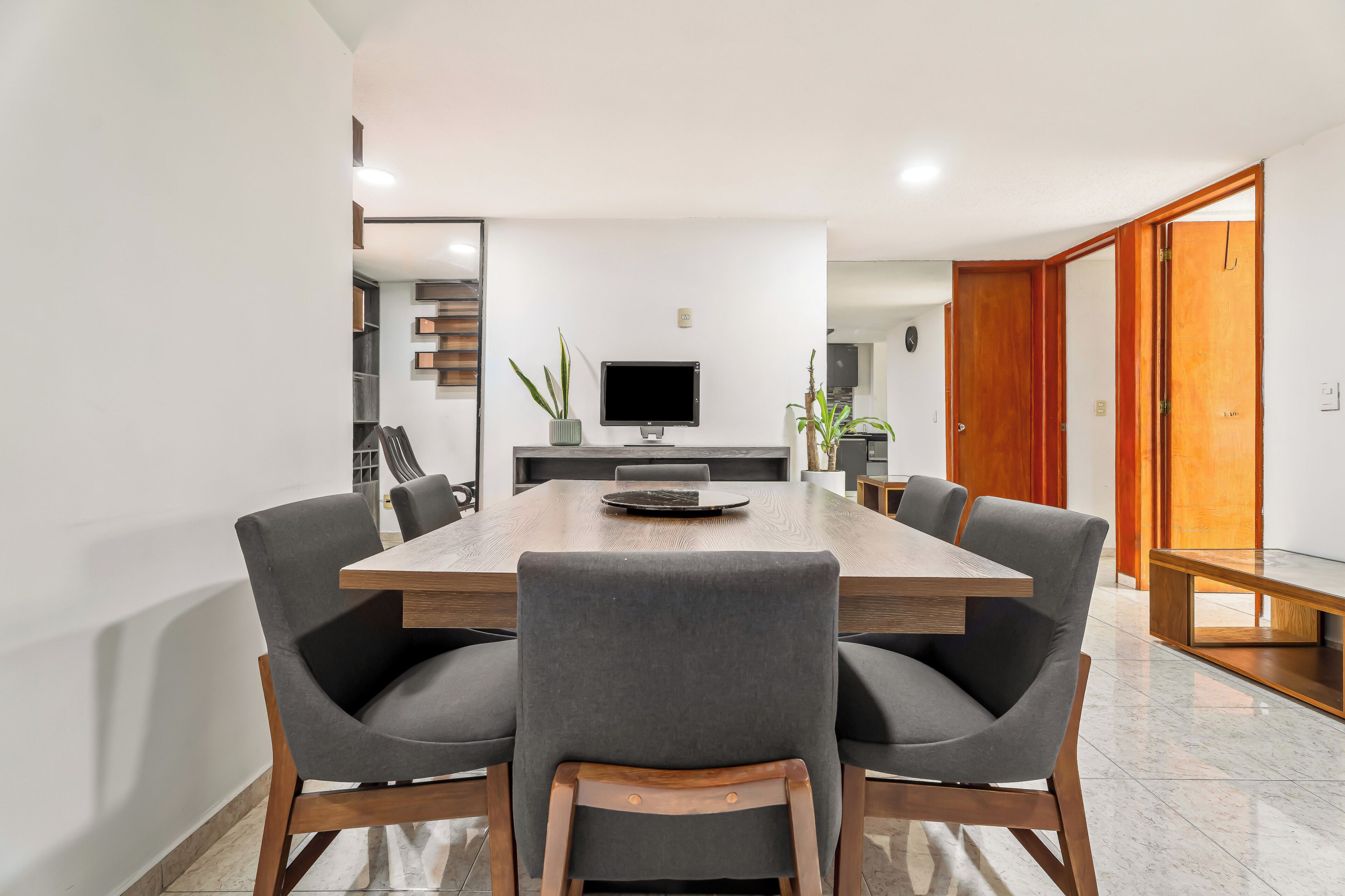 Un comedor moderno con una larga mesa de madera, seis sillas grises tapizadas, un televisor de pantalla plana y una planta en maceta.