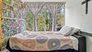 Ein Schlafzimmer mit einem Bett, einem Bücherregal, einem Fenster mit Vorhängen und einem Kreuz an der Wand.